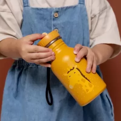 Garrafa amarela com estampa de girafa nas mãos de criança com roupa azul e bege, fundo castanho.