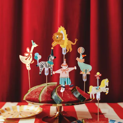Pedaços de melancia com espetos decorativos de papel de animais de circo sobre uma mesa com toalha listrada