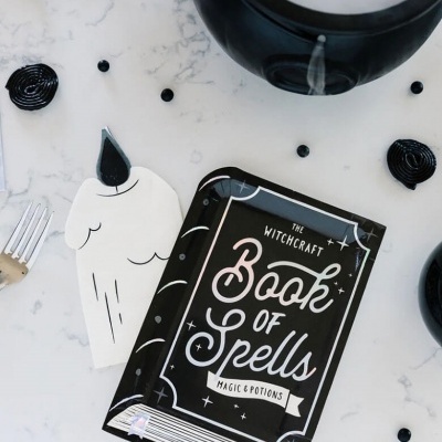 Livro de feitiços e caixa em forma de vela sobre mesa marmoreada com doces pretos.