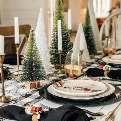 Mesa decorada com pratos, guardanapos e árvores de Natal artificiais em tons de verde e branco.