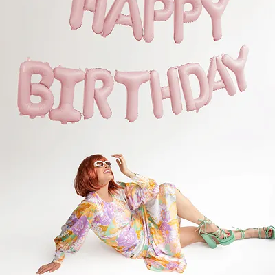 Balões Happy Birthday cor-de-rosa com mulher em vestido florido sentada no chão