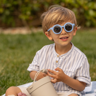 Criança com óculos de sol azuis e camisa riscada a segurar balde bege no jardim.