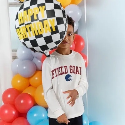 Menino com camisola branca e calças pretas segura balão 'Happy Birthday' em festa com balões coloridos