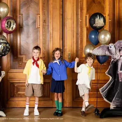 Crianças com roupas coloridas e balões junto a figuras de Harry Potter e Dobby em ambiente com porta de madeira.
