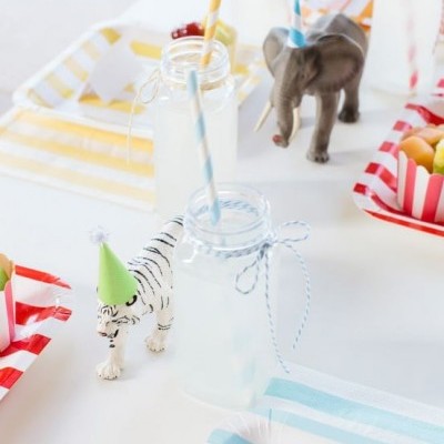 Frascos transparentes com canudos e figuras de animais sobre mesa com pratos de papel às riscas