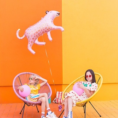 Crianças sentadas em cadeiras coloridas segurando balões cor-de-rosa em fundo laranja e amarelo