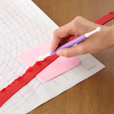 Ferramenta roxa para modelar pasta de açúcar vermelha numa superfície rosa e placa branca com marcações