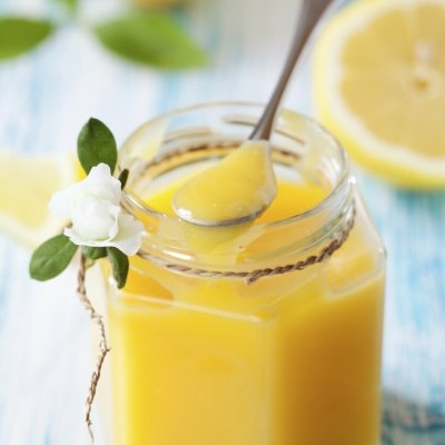 Frasco de vidro com creme amarelo e colher, decorado com flor, limões ao fundo.