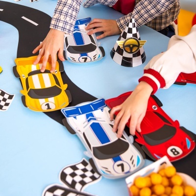 Carros de corrida de papel coloridos em mesa com pista e chapéu de festa às riscas