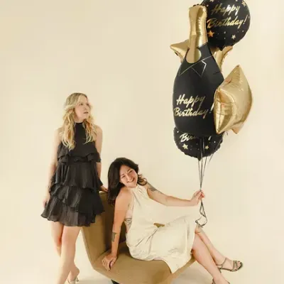Duas mulheres com vestidos elegantes e balões de Happy Birthday num fundo neutro.