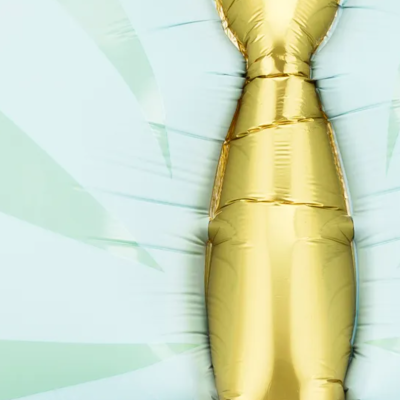 Balão metálico dourado em forma de gravata sobre superfície clara com sombras verdes