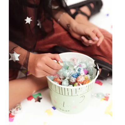 Balde verde claro com rebuçados coloridos e pessoa sentada vestindo capa preta com estrelas e lua, em chão branco com confetes.