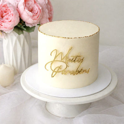 Bolo branco redondo com texto dourado Muitos Parabéns e pétalas cor-de-rosa ao lado