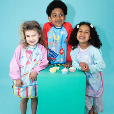 Três crianças com aventais coloridos e cupcakes sobre bloco verde em fundo azul