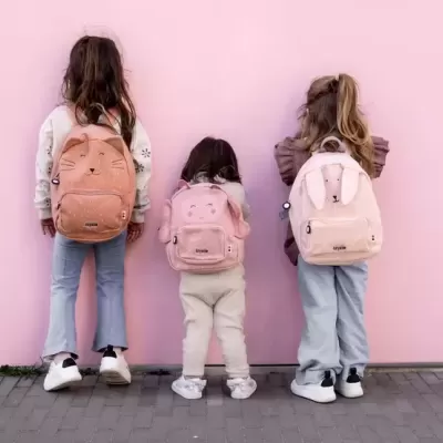 Três crianças com mochilas rosa de peluche com carinhas de animais numa parede rosa