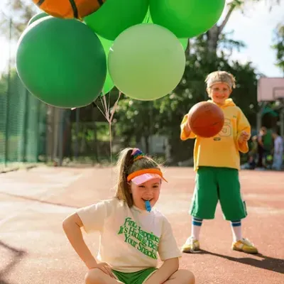 Duas crianças num campo de basquetebol com balões, uma sentada com t-shirt e calções verdes, outra com roupa amarela e calções verdes a jogar basquetebol