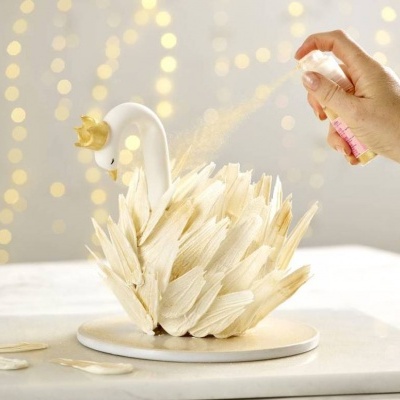 Cisne decorativo branco com coroa dourada e base circular, sendo pulverizado com spray por uma mão