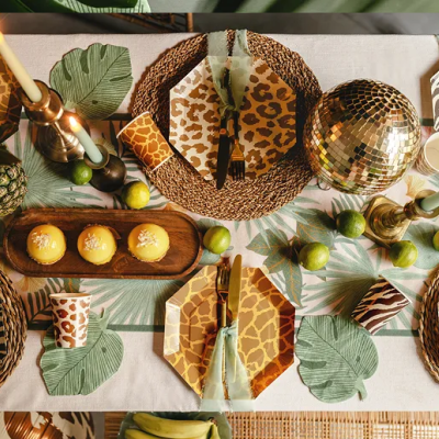 Mesa posta com tema de animais com pratos de papel, guardanapos verdes, velas, frutas tropicais, bolos amarelos, copos com padrão de pele de leopardo e disco de espelhos