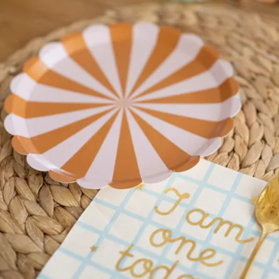 Prato descartável branco e laranja com guardanapo decorativo e talher dourado num suporte trançado