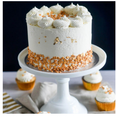 Bolo branco decorado com confeitos dourados num suporte branco, rodeado por cupcakes