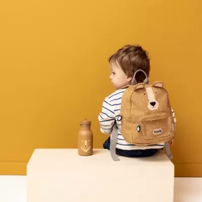 Criança com mochila castanha de urso sentada numa caixa clara e garrafa ao lado