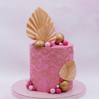 Bolo rosa decorado com folhas douradas secas e bolas coloridas, sobre base redonda rosa