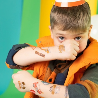 Criança com tatuagens temporárias de carros e sinais em braços, vestindo colete laranja e camisa azul e laranja.