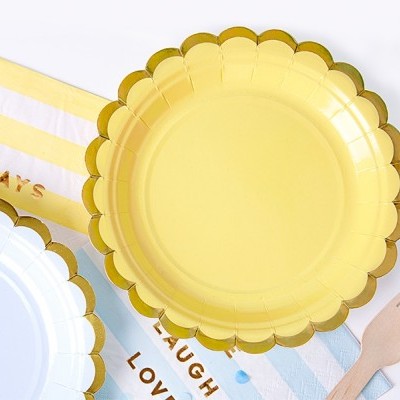 Pratos descartáveis amarelo e branco com bordas douradas, toalhas de papel com texto e garfo de madeira