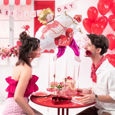 Casal numa mesa vermelha decorada com balões vermelhos e de unicórnio 'LOVE', decoração de Dia dos Namorados.