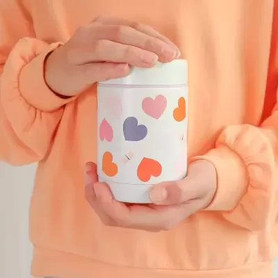Frasco branco decorado com corações coloridos e pequenas borboletas, mãos segurando frasco, camisola cor de pêssego