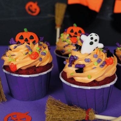 Cupcakes decorados com tema de Halloween, com abóboras e fantasma, em formas roxas e mini vassouras no fundo