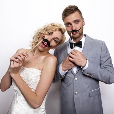 Casal de noivos vestidos a rigor com bigodes falsos em pauzinhos.