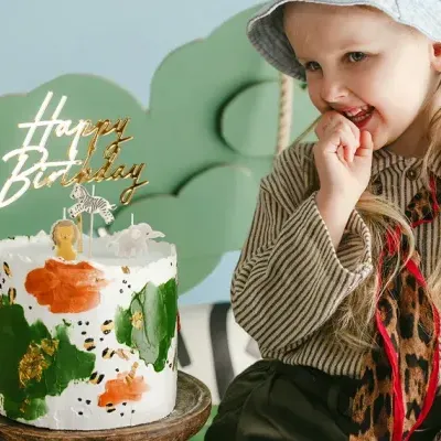 Bolo de aniversário decorado com glacê branco e manchas verdes e laranja com topo dourado Happy Birthday ao lado de criança loira com chapéu