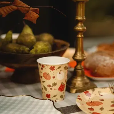 Copo de papel com padrão de outono, pratos e castiçal dourado numa mesa com pêras