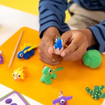 Figuras de dinossauros de plasticina colorida em fundo laranja com mãos de criança segurando uma peça