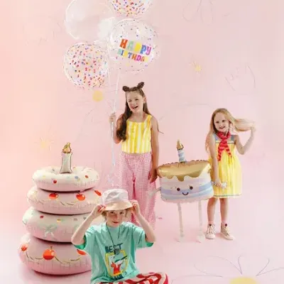Crianças com balões de aniversário, boias em forma de donuts e puff em forma de bolo num fundo rosa com desenhos de flores.