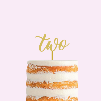 Bolo de camadas com cobertura branca semi-nua e topper dourado 'two' sobre fundo rosa claro