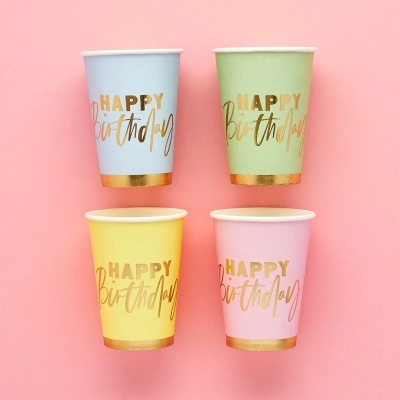 Quatro copos de papel festivos coloridos com texto dourado HAPPY birthday em fundo rosa