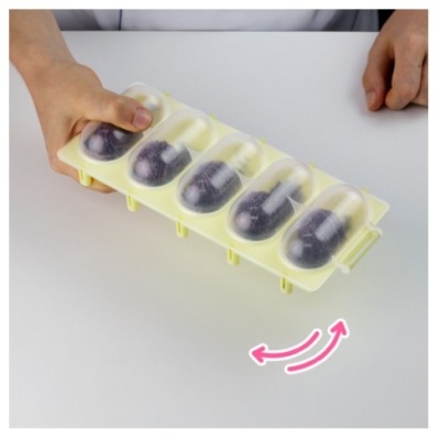 Forma de gelo amarela com bolas de azeitona pretas dentro, com tampa transparente, sobre superfície branca