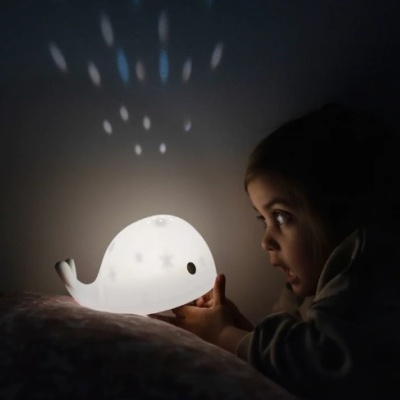 Lâmpada decorativa baleia branca com projeção de estrelas e criança a observar.