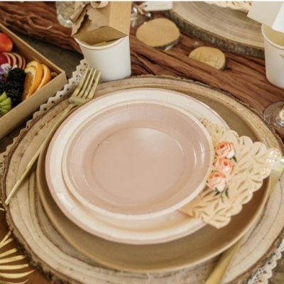 Conjunto de pratos rosa claro com talheres dourados e guardanapo branco decorado, sobre base de madeira