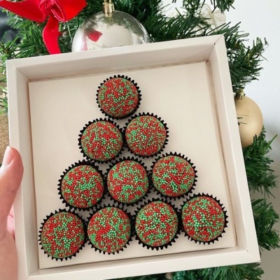 Conjunto de brigadeiros decorados dispostos em forma de árvore de Natal numa caixa branca em frente a uma árvore de Natal decorada