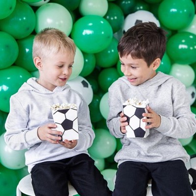 Dois rapazes segurando caixinhas de pipocas com padrão de bola de futebol, rodeados por balões verdes, brancos e pretos.