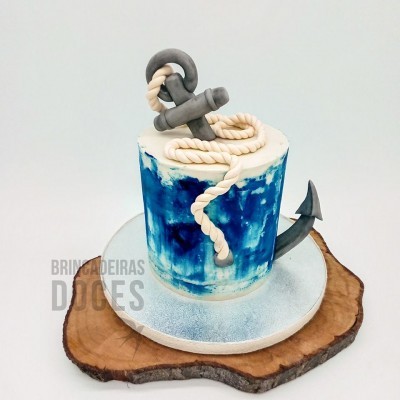 Bolo azul e branco com âncora e corda decorativos sobre prato prateado e base de madeira
