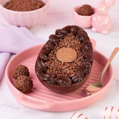 Ovo de chocolate recheado com granulados e decoração de chocolate preto num prato rosa com duas trufas e colher dourada