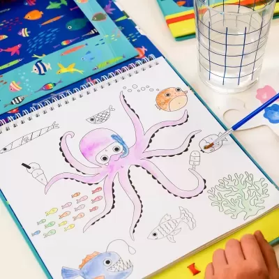 Caderno de colorir com desenho de polvo e peixes, pincel e copo de água