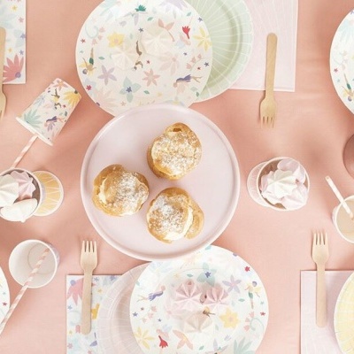 Mesa posta com pratos descartáveis florais, copos rosa e pães num prato branco