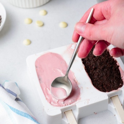 Molde branco de plástico com mistura rosa para gelado e base castanha escura, mão segurando colher, recipientes com pepitas de chocolate ao fundo.