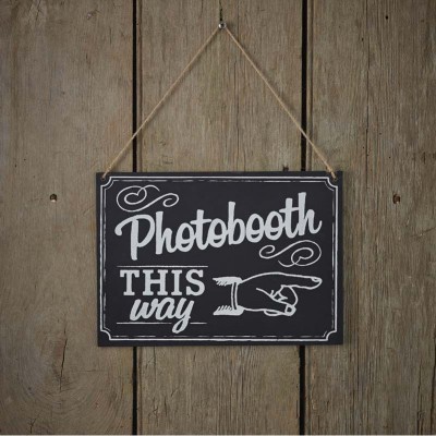 Placa decorativa preta com texto branco 'Photobooth THIS way' pendurada num fundo de madeira