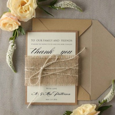 Convite decorativo com envelope castanho, papel branco com texto e faixa de juta, rodeado por flores creme e verdes sobre fundo cinza